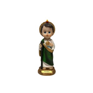 May include: A small figurine of Saint Jude Thaddeus, a Catholic saint, depicted as a young boy with a halo and a staff. He is wearing a green robe and a white tunic. The figurine is made of resin and has a gold base with the inscription "San Judas".