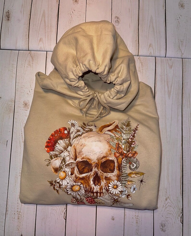 May include: A beige hoodie with a skull and floral design. The skull is surrounded by flowers, mushrooms, and leaves. The hoodie has a drawstring hood.