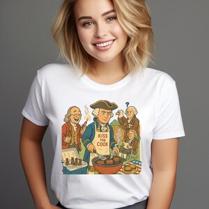 May include: White t-shirt with a cartoon illustration of historical figures at a barbecue. The central figure wears an apron that says "Kiss the Cook." Other figures are shown with burgers, beer, and fireworks. The design is in a cartoon style.