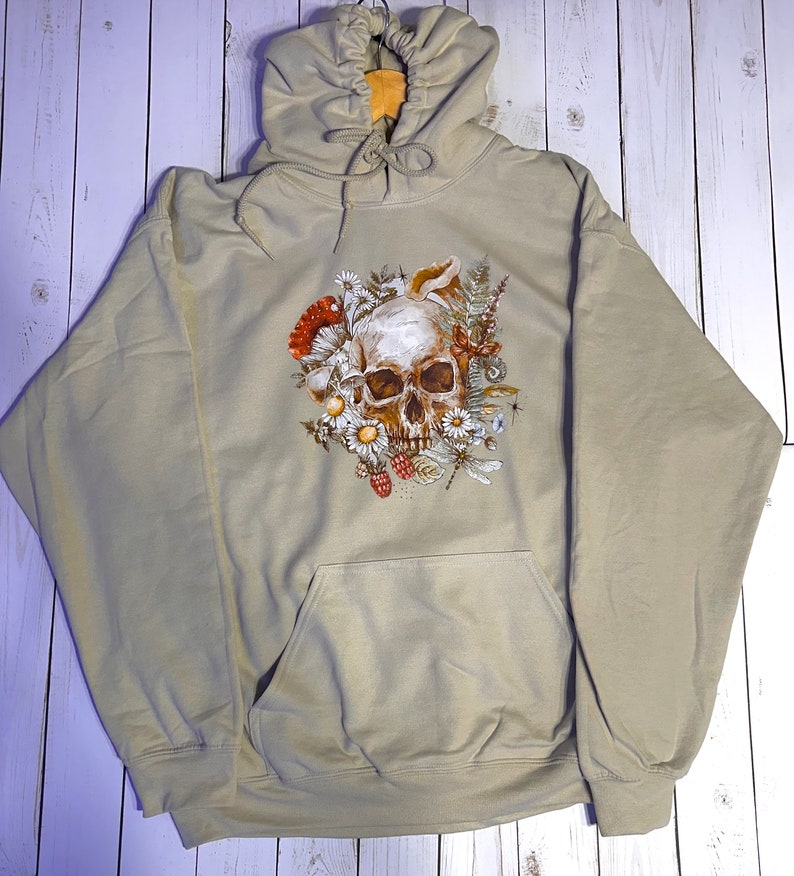May include: A beige hooded sweatshirt with a skull surrounded by flowers, mushrooms, and insects printed on the front.