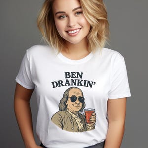 May include: White t-shirt with the text "BEN DRANKIN'" above an illustration of Benjamin Franklin wearing sunglasses and holding a red cup. The graphic is in a cartoon style.
