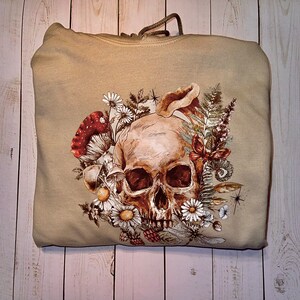 May include: A beige hoodie with a skull and floral design. The skull is surrounded by flowers, leaves, and mushrooms. The design is in shades of brown, green, and red.