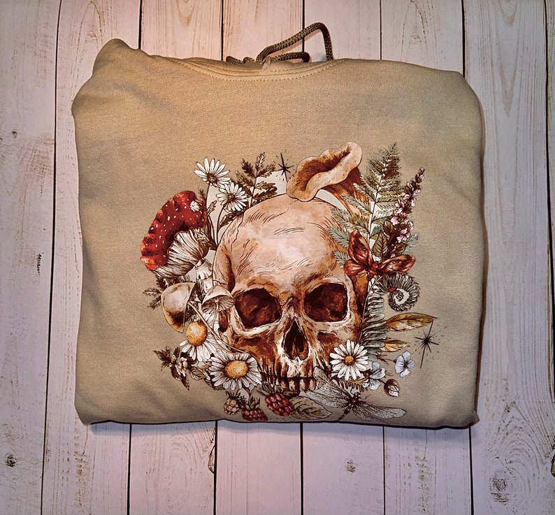 May include: A beige hoodie with a skull and floral design. The skull is surrounded by flowers, leaves, and mushrooms. The design is in shades of brown, green, and red.