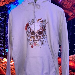 May include: A light gray hoodie with a skull and floral design. The skull is surrounded by flowers, leaves, and berries. The design is in shades of brown, white, and green.