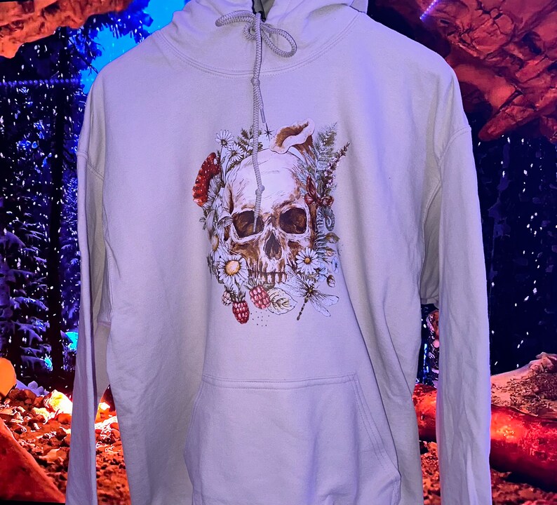 May include: A light gray hoodie with a skull and floral design. The skull is surrounded by flowers, leaves, and berries. The design is in shades of brown, white, and green.