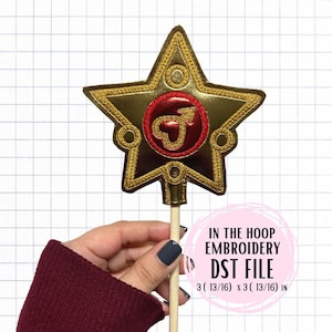 May include: Gold star-shaped embroidery design with a red circle in the center featuring a stylized arrow. The design is on a wooden stick and measures 3 13/16 inches by 3 13/16 inches. IN THE HOOP EMBROIDERY DST FILE.