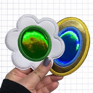 May include: Two embroidered patches: one flower-shaped, the other oval. The flower patch is white with a green center and silver trim. The oval patch has a gold border, a blue center, and a blue trim. Both patches have a reflective, iridescent quality.