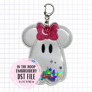 Peut inclure: Un porte-clés blanc en forme de fantôme avec un nœud rose et des yeux noirs. Le porte-clés a une bordure argentée et une poche transparente remplie d'étoiles et de confettis colorés. Le texte "IN THE HOOP EMBROIDERY DST FILE 7.6 x 7.6 (2.7) cm" est dans un cercle rose.