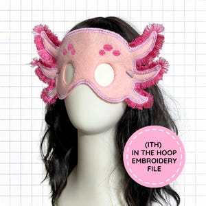 May include: A light pink felt mask shaped like an axolotl with dark pink embroidered fringe gills and eye holes. The mask features small dark pink embroidered spots above the eyes. A circular pink label reads (ITH) IN THE HOOP EMBROIDERY FILE.