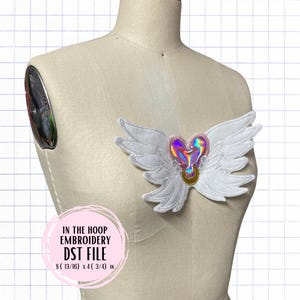 May include: White wings with a holographic pink heart in the center. The wings are on a white mannequin. The text "IN THE HOOP EMBROIDERY DST FILE 8 (13/16) x 4 (3/4) IN" is below the wings.