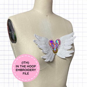 May include: An embroidered patch featuring white wings surrounding a central iridescent pink heart with a gold crescent moon accent. A pink circular graphic with the text (ITH) IN THE HOOP EMBROIDERY FILE is overlaid on the lower left side.