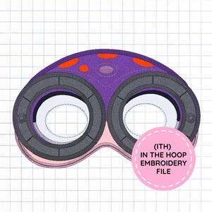 May include: A purple eye mask with orange spots and two dark gray circular eye frames. The mask features a light pink lower section and includes a pink circular badge with the text (ITH) IN THE HOOP EMBROIDERY FILE printed in black.