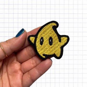 Peut inclure: Un patch brodé en forme d'étoile jaune avec des contours noirs et deux yeux noirs. Le patch est tenu dans une main.