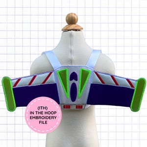 Peut inclure: Un accessoire jetpack en tissu blanc, violet et vert citron avec des rayures diagonales rouges sur les ailes, un ovale violet central et une étiquette circulaire rose avec le texte (ITH) IN THE HOOP EMBROIDERY FILE sur un fond quadrillé.