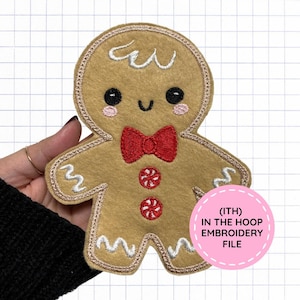 May include: A tan felt gingerbread person applique featuring embroidered black eyes, pink cheeks, a red bow tie, and two red and white peppermint buttons. White icing details decorate the head, arms, and legs. Text reads (ITH) IN THE HOOP EMBROIDERY FILE.