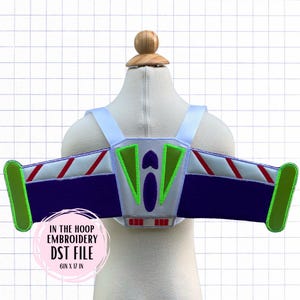 Puede incluir: Accesorio de disfraz con temática de Buzz Lightyear, con un diseño de alas blancas, verdes, rojas y azules. Las alas están unidas a correas blancas. El texto "IN THE HOOP EMBROIDERY DST FILE 6IN X 17 IN" es visible.