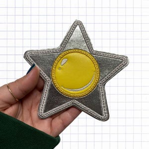 Peut inclure: Un écusson en forme d'étoile argentée avec un cercle jaune au centre. Le cercle a un contour blanc et un petit cercle blanc à l'intérieur.