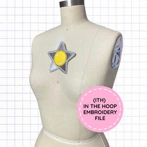 May include: A beige dress form displays a silver star-shaped patch with a central yellow circle. A pink circular graphic on the lower right contains the text (ITH) IN THE HOOP EMBROIDERY FILE. A second patch is visible on the shoulder area.