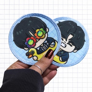 May include: Two circular embroidered patches featuring a cartoon character with black hair. One patch shows the character wearing rainbow flower-shaped glasses and a yellow duck floatie. The other patch shows the character with a frustrated expression.