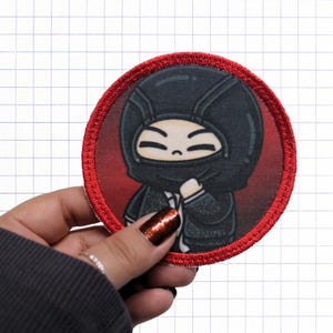 Puede incluir: Un parche bordado circular que muestra un personaje de dibujos animados con un pasamontañas negro y una chaqueta oscura sobre un fondo rojo intenso. El parche tiene un borde cosido de color rojo vibrante y una superficie impresa lisa.