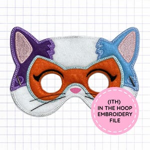May include: A cat-shaped felt mask featuring a white base, purple and blue ears, and an orange eye patch area. The mask includes black embroidered eyelashes and whiskers, a pink nose, and a circular pink label with the text (ITH) IN THE HOOP EMBROIDERY FILE.