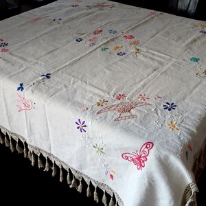 May include: White linen tablecloth with embroidered floral designs, birds, and a butterfly. The tablecloth has a crocheted edge with tassels.