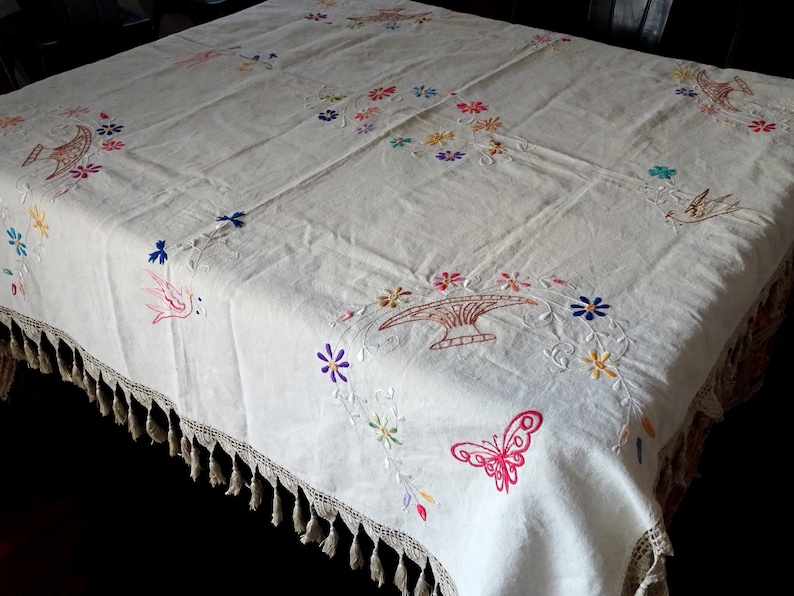 May include: White linen tablecloth with embroidered floral designs, birds, and a butterfly. The tablecloth has a crocheted edge with tassels.
