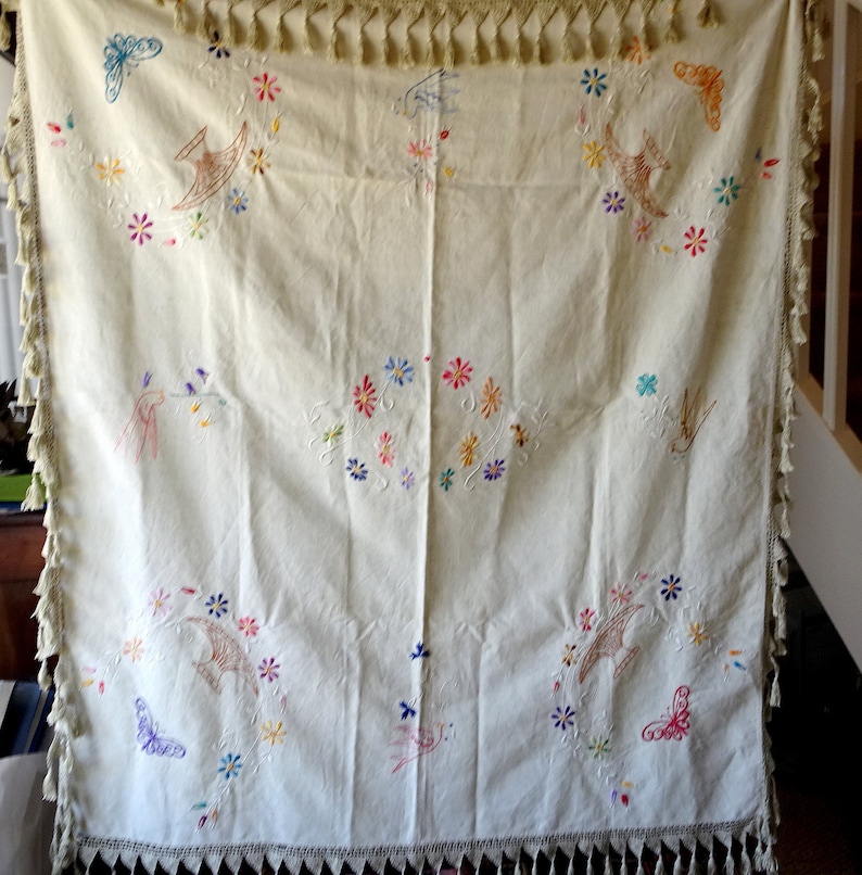 May include: A white tablecloth with embroidered birds, butterflies and flowers in various colours. The tablecloth has a fringed border.