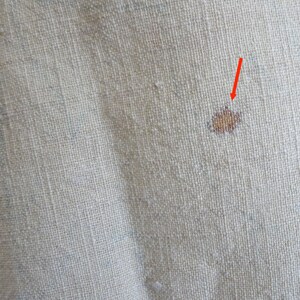 May include: Close-up of a beige fabric with a small brown stain. The stain is in the centre of the image and is surrounded by a light beige fabric.