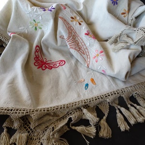 May include: A white linen tablecloth with embroidered flowers, butterflies, and a brown design. The tablecloth has a crocheted border with tassels.