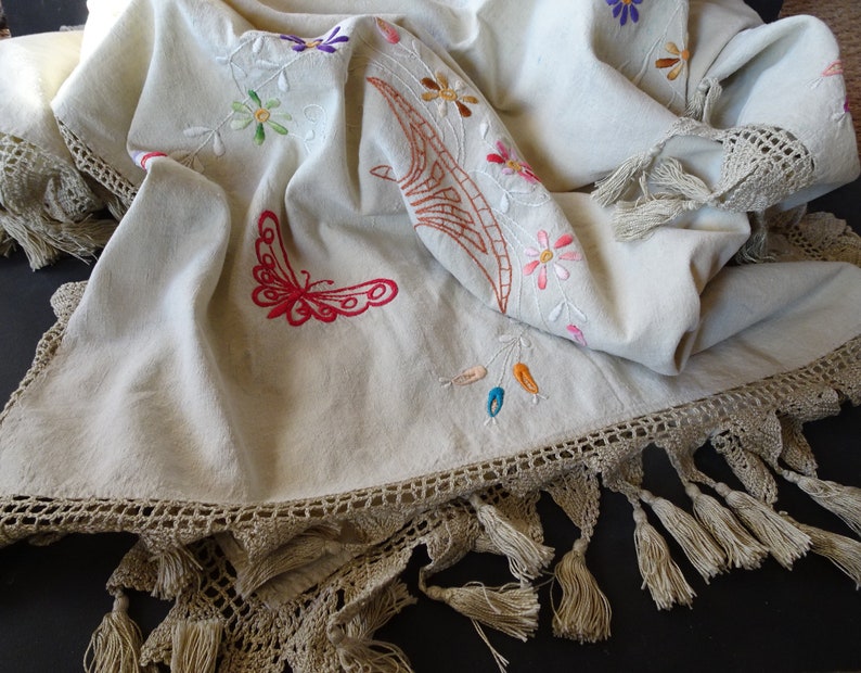 May include: A white linen tablecloth with embroidered flowers, butterflies, and a brown design. The tablecloth has a crocheted border with tassels.