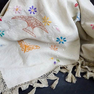 May include: A white linen tablecloth with a crocheted border and tassels. The tablecloth is embroidered with colourful flowers, a butterfly, and a stylised design.