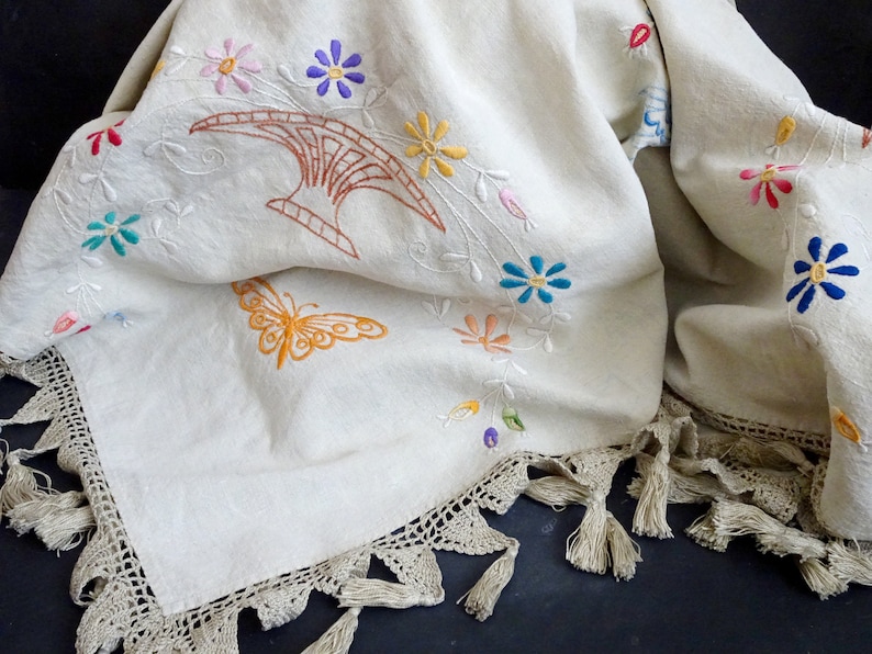 May include: A white linen tablecloth with a crocheted border and tassels. The tablecloth is embroidered with colourful flowers, a butterfly, and a stylised design.