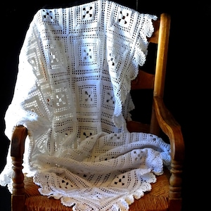 May include: A white, crocheted blanket draped over a wooden chair. The blanket features a square pattern with openwork details and a scalloped edge. The chair has a woven seat and a dark wood finish, set against a black background.