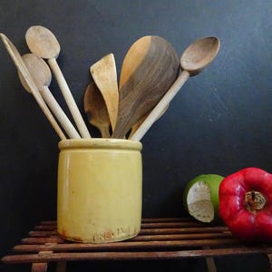 Puede incluir: Un tarro de cerámica amarillo con una colección de cucharas y espátulas de madera en su interior. El tarro está sentado en un estante de madera.