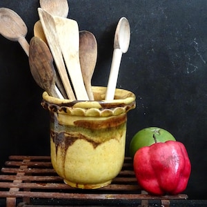 Peut inclure: Un pot à ustensiles en céramique jaune avec un glaçage brun et un bord festonné, rempli de cuillères et de spatules en bois. Le pot est posé sur une grille métallique avec un poivron vert et rouge au premier plan.