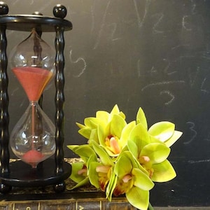 May include: A black hourglass with red sand, alongside a bouquet of green and yellow flowers. The hourglass is made of glass and wood. The background is a chalkboard with white markings. The flowers are in the foreground.