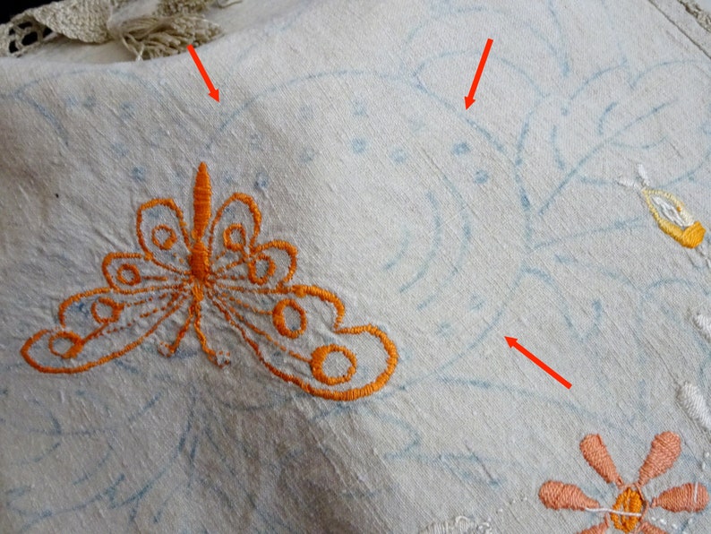 May include: A white linen tablecloth with embroidered details. The embroidery includes a butterfly, flowers, and leaves. The embroidery is done in orange, blue, and pink thread.