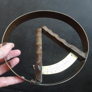 Könnte beinhalten: Ein Messwerkzeug aus Messing und Holz mit einem kreisförmigen Band und einem Schieberegler. Der Schieberegler ist in Zoll unterteilt.