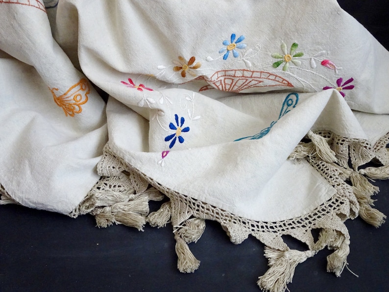 May include: A white linen tablecloth with embroidered flowers and butterflies. The tablecloth has a crocheted edge with tassels.