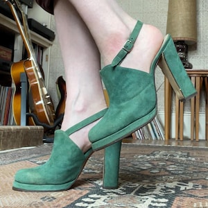 May include: A pair of green suede heeled sandals with an ankle strap. The sandals have a pointed toe and a thick heel.