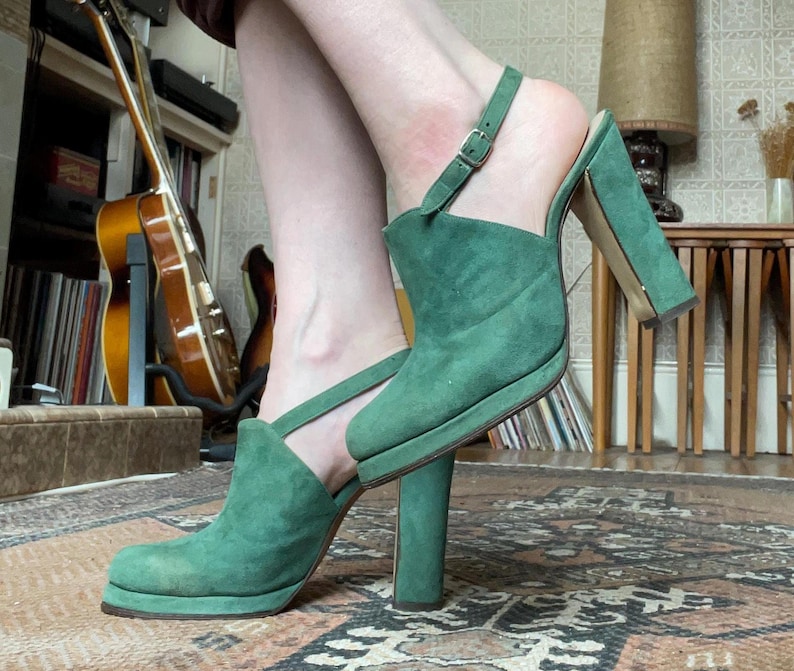May include: A pair of green suede heeled sandals with an ankle strap. The sandals have a pointed toe and a thick heel.