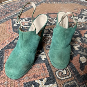May include: A pair of green suede heeled shoes with a slingback strap. The shoes have a chunky heel and a rounded toe.