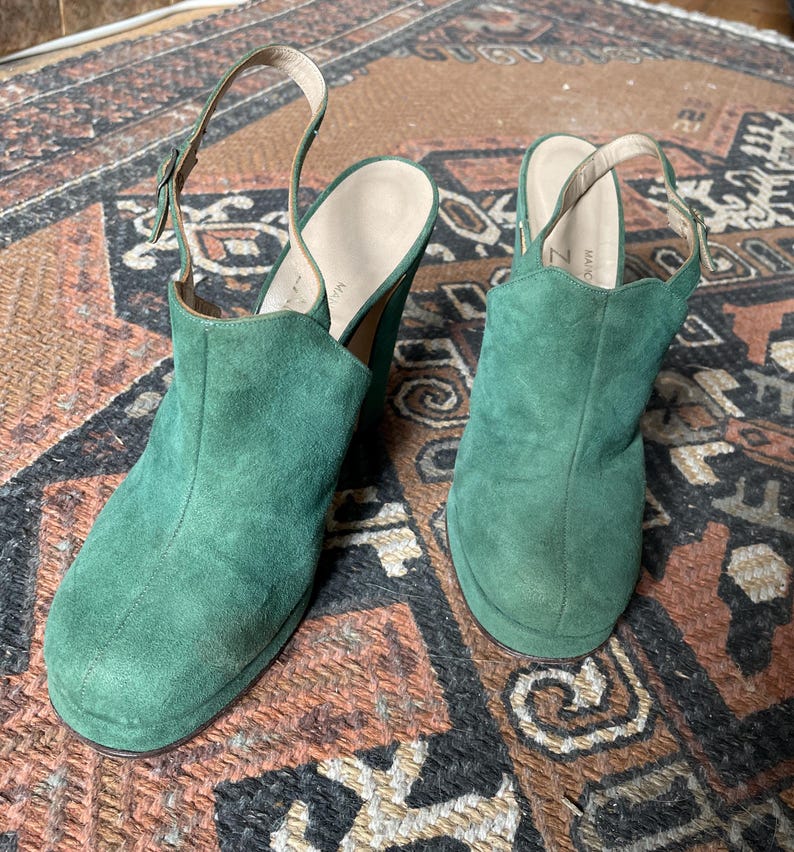 May include: A pair of green suede heeled shoes with a slingback strap. The shoes have a chunky heel and a rounded toe.