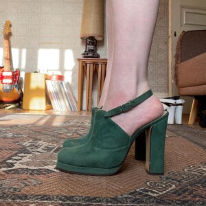 May include: Green suede heeled shoes with a strap and buckle. The shoes have a platform sole and a chunky heel.