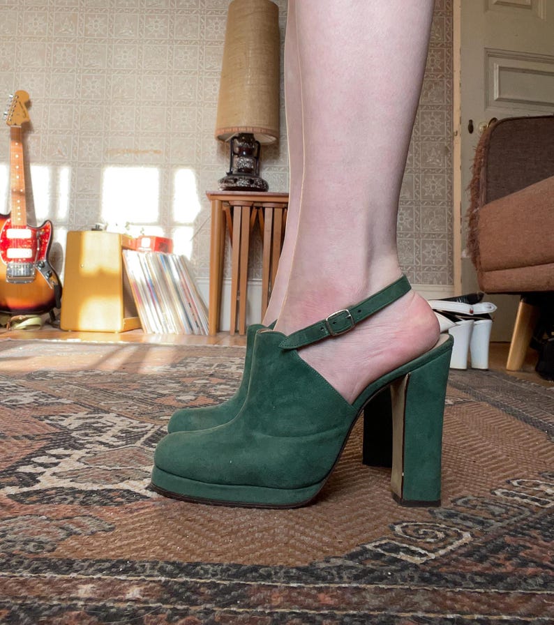 May include: Green suede heeled shoes with a strap and buckle. The shoes have a platform sole and a chunky heel.