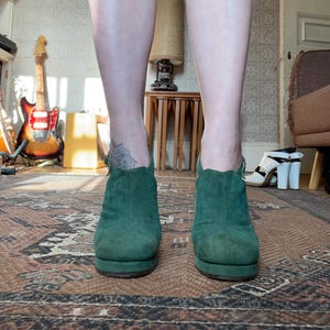 May include: A pair of dark green suede ankle boots with a chunky heel. The boots have a pointed toe and a zipper on the back.