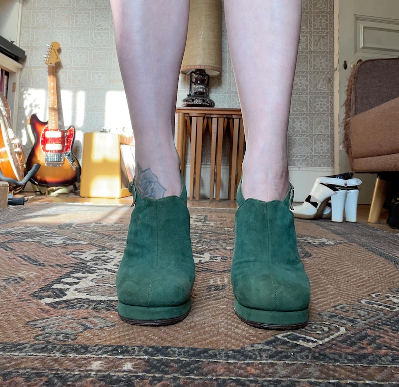 May include: A pair of dark green suede ankle boots with a chunky heel. The boots have a pointed toe and a zipper on the back.