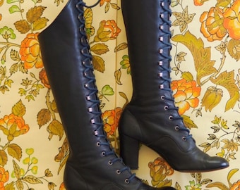May include: A pair of black leather knee-high boots with lace-up fronts and chunky heels. The boots are on a floral patterned background.