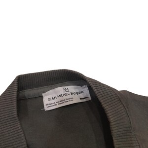 May include: Close-up of a dark olive green sweatshirt collar. A white tag displays the text "JEAN MICHEL BASQUIAT" with a crown logo. The tag also includes licensing information from Artestar, New York.